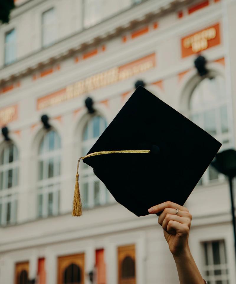 Graduate celebrating success — holding a graduation cap in front of a university building